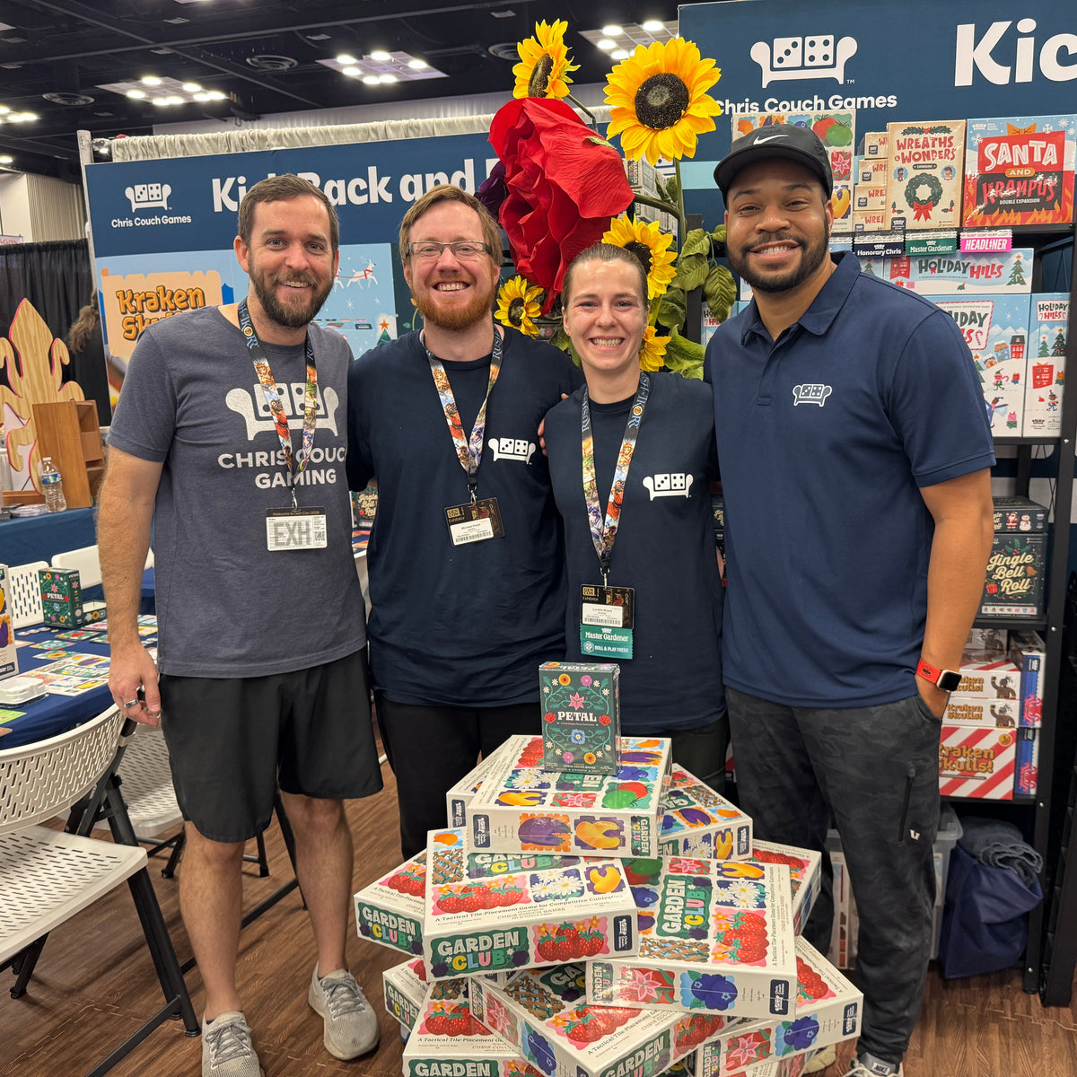 Four people standing together at a convention booth surrounded by game boxes and displays. Two men wear matching navy blue shirts with 'Kick Back and Play' printed on the back, and a stack of colorful game boxes sits in front of them.